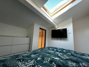 Ein Schlafzimmer mit Dachfenster, einem Bett mit gemustertem Überwurf, einem an der Wand befestigten Fernseher und einem Kleiderschrank.