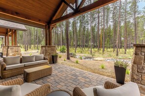 A covered outdoor seating area with stone pillars, wicker furniture, and a view of a forest. - NEW! 5 BR Luxury Home, Sleeps 12 - ELKR56 (Sunriver)