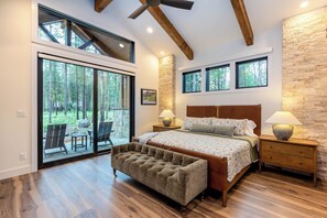 Quarto com cama grande, cabeceira de madeira, duas mesas de cabeceira com luminárias, um banco e uma porta de vidro deslizante que dá para uma área externa com cadeiras.