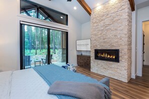 Ein Schlafzimmer mit einem großen Bett, einem Kamin aus Stein, Blick auf einen Wald durch eine Glastür und einem an der Wand montierten Fernseher.