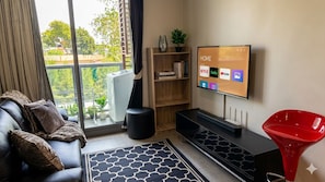 Un salon équipé d’un téléviseur diffusant des services de streaming, d’un canapé, d’une table basse, d’une bibliothèque et d’une fenêtre donnant sur un paysage agréable.