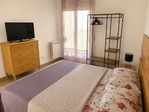 A bedroom with a bed, a wooden dresser, a TV on top, a radiator, a window with curtains, a metal shelf, and a lamp on a side table.