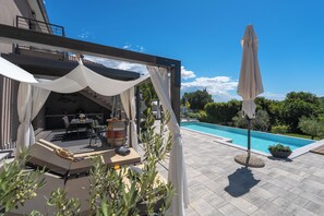 Ett takförsett utomhusområde med sittplatser, och en pool syns i bakgrunden.