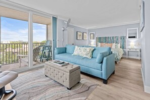 Ein modernes Wohnzimmer mit einem blauen Sofa, einem passenden Hocker, einem großen Fenster und einem Balkon.