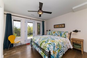 Une chambre à coucher avec un lit, un ventilateur de plafond, une chaise, une table de chevet et une vue sur l’extérieur, visible à travers la porte.