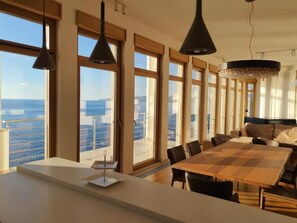 Ett rymligt matområde med ett stort bord och stolar, stora fönster med utsikt över havet och hängande lampor.