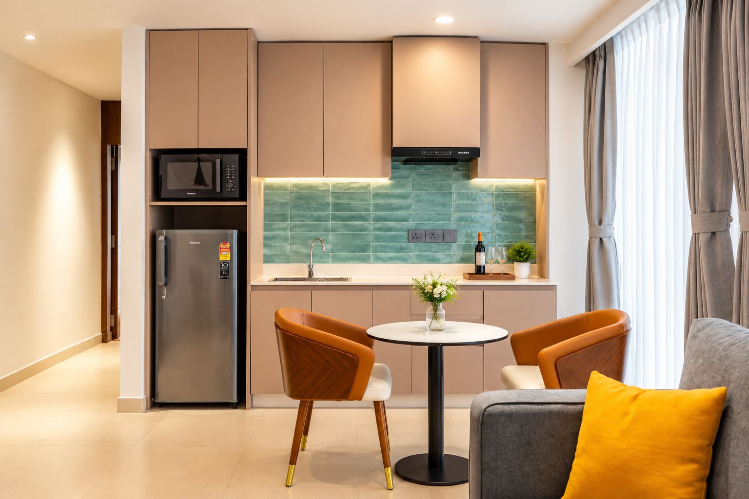 Cocina moderna con microondas integrado, refrigerador y salpicadero verde. Cuenta con una zona de comedor con dos sillas y una mesa pequeña.