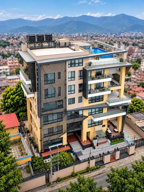 Un edificio de varios pisos con balcones, piscina en la azotea y vista a un paisaje urbano con montañas al fondo.
