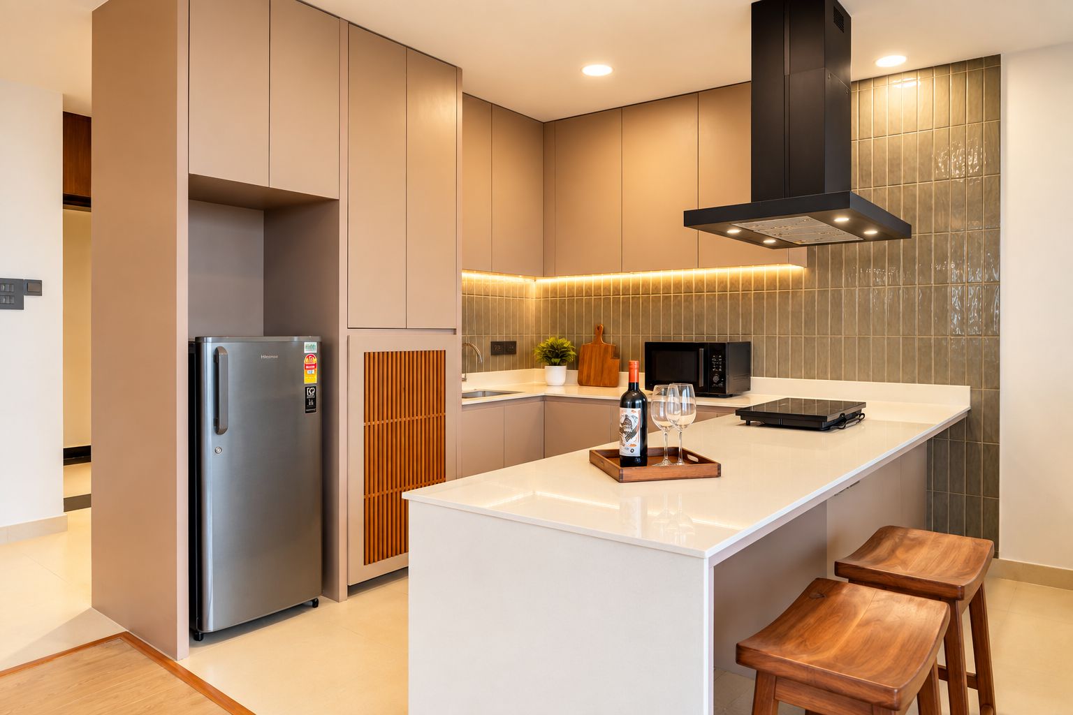 Cozinha moderna com geladeira de aço inoxidável, micro-ondas e forno embutido. Há uma ilha de madeira com banquetas e uma garrafa de vinho.
