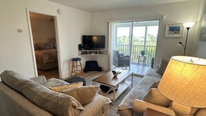 En stue med sofa, sofabord, lampe, fjernsyn og en balkon med udsigt.