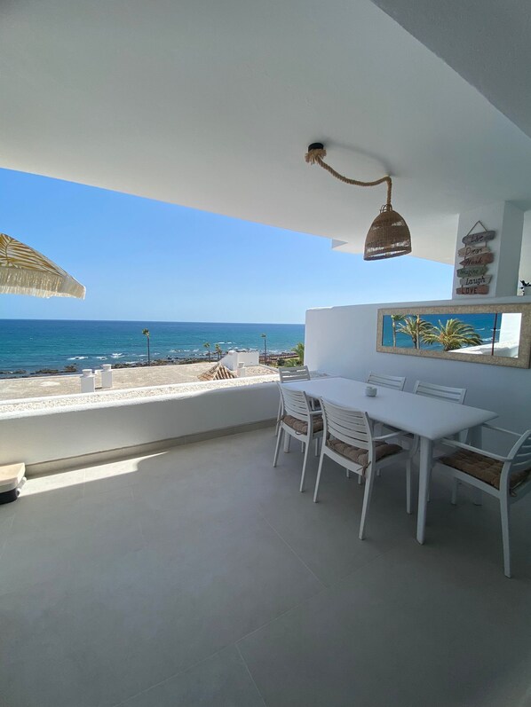 Ein Balkon mit weißem Tisch und Stühlen, einer Hängeleuchte und Blick aufs Meer.
