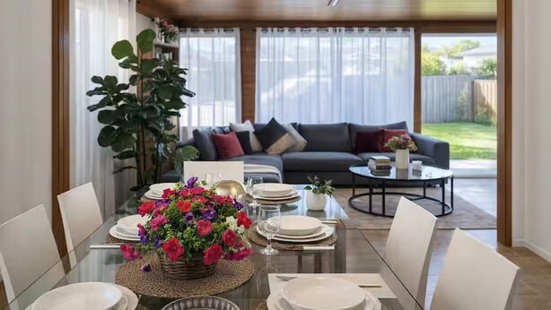 Un comedor con una mesa de vidrio, sillas blancas y un centro de mesa floral. Se ve un sofá y una mesa de centro al fondo.