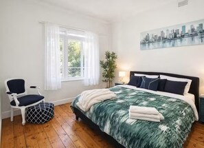 A bedroom with a bed, a chair, a window with curtains, a plant, and a cityscape painting on the wall. - Spacious 4-bed Retreat in Vibrant Footscray (Melbourne)