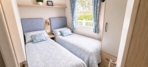 Une chambre à coucher de taille modeste, avec deux lits, une petite étagère ornée d’une plante et d’un tableau, et une fenêtre avec des rideaux.