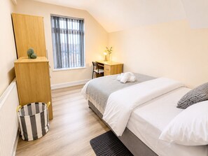 A bedroom with a bed, a desk, a chair, a window with curtains, a radiator, and a storage unit. - The Shed - Cosy 2-Bed Flat with Free Parking - Close to City Centre (Cardiff)