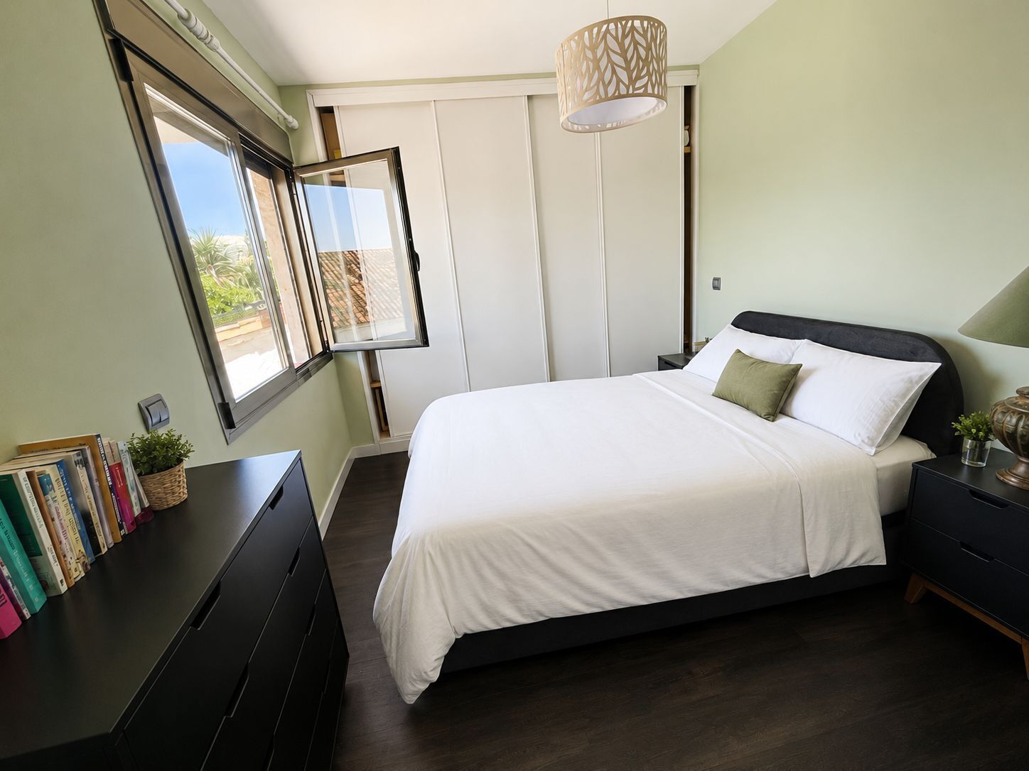 Une chambre à coucher avec un lit, des tables de chevet, un bureau avec des livres, une fenêtre et un luminaire suspendu.