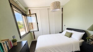 Une chambre à coucher avec un lit, des tables de chevet, un bureau avec des livres, une fenêtre et un luminaire suspendu.