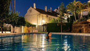 Une piscine avec une personne dans l’eau, entourée de palmiers et d’un bâtiment doté d’un toit en tuiles.