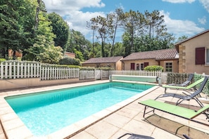 Une piscine avec des chaises longues, une clôture blanche en bois et un bâtiment doté d’un toit en tuiles.