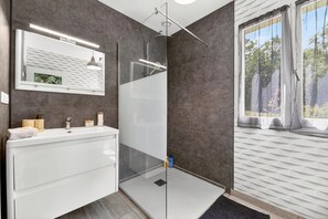 A modern bathroom with a glass shower enclosure, a white vanity, and a window with a view of trees.