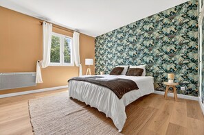 A bedroom with a bed, a window with curtains, a radiator, a patterned wallpaper, and a small wooden stool.
