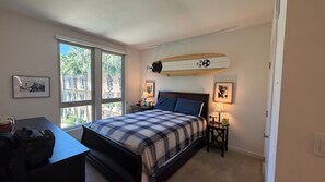 Une chambre avec un lit, un bureau, une décoration sur le thème du surf et deux fenêtres.