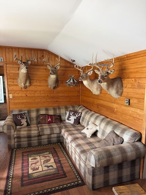 A cozy cabin interior with a wooden ceiling, a plaid sofa, and mounted deer heads.