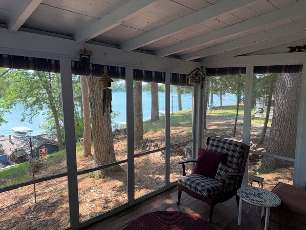 A covered porch with a view of a lake, a chair, a small table, and a hanging bird feeder.