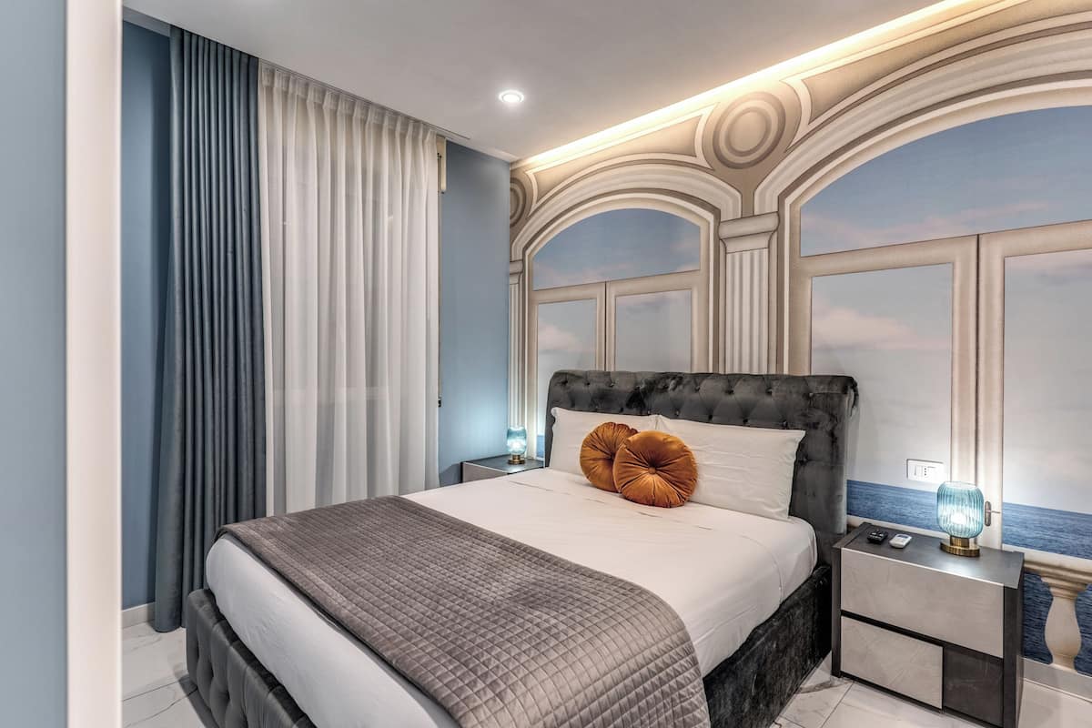 Une chambre à coucher avec un grand lit, une table de chevet, une lampe et une vue sur la mer par une fenêtre.