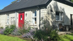 A house with a red door, a sloped roof, and a garden with purple flowers. - Cosy 2 bedroom  holiday apartment in a beautiful quiet area beside a lake. (Bantry)