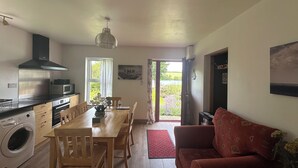 A kitchen with a microwave, stove, and washing machine; a dining table with chairs; a red sofa; and a door leading outside. - Cosy 2 bedroom  holiday apartment in a beautiful quiet area beside a lake. (Bantry)