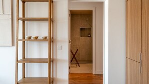 Un couloir avec une étagère en bois, une porte donnant sur une pièce dont le sol est recouvert de carreaux, et une armoire en bois.