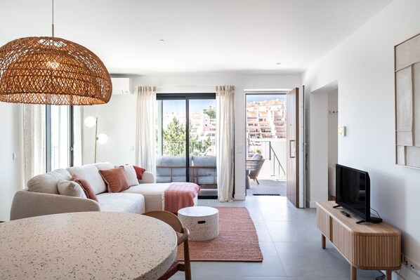 Ein modernes Wohnzimmer mit einer Couch, einem Esstisch, einem Fernseher und einem Balkon mit Blick auf Gebäude.