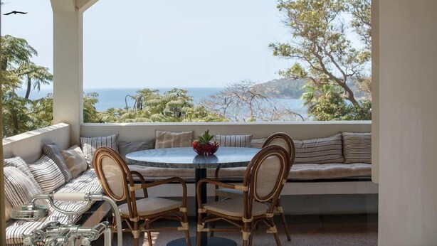 A covered outdoor seating area with a round table and chairs, overlooking a scenic view of the sea and trees. - Le Séku bi (Dakar)