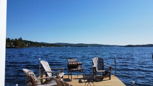 A wooden deck with two Adirondack chairs and a table, overlooking a calm lake with a distant shoreline. - STAY ABOVE THE WATER ON BEAUTIFUL LAKE WINNIPESAUKEE (Laconia)