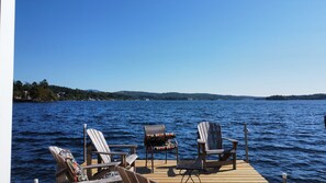 Terraza de madera con dos sillas Adirondack y una mesa, con vista a un lago tranquilo y una orilla lejana.