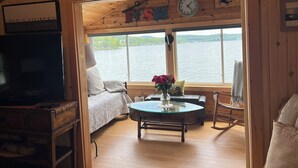 A cabin interior with a bed, a table with a vase of flowers, a clock, and a view of a lake. - STAY ABOVE THE WATER ON BEAUTIFUL LAKE WINNIPESAUKEE (Laconia)