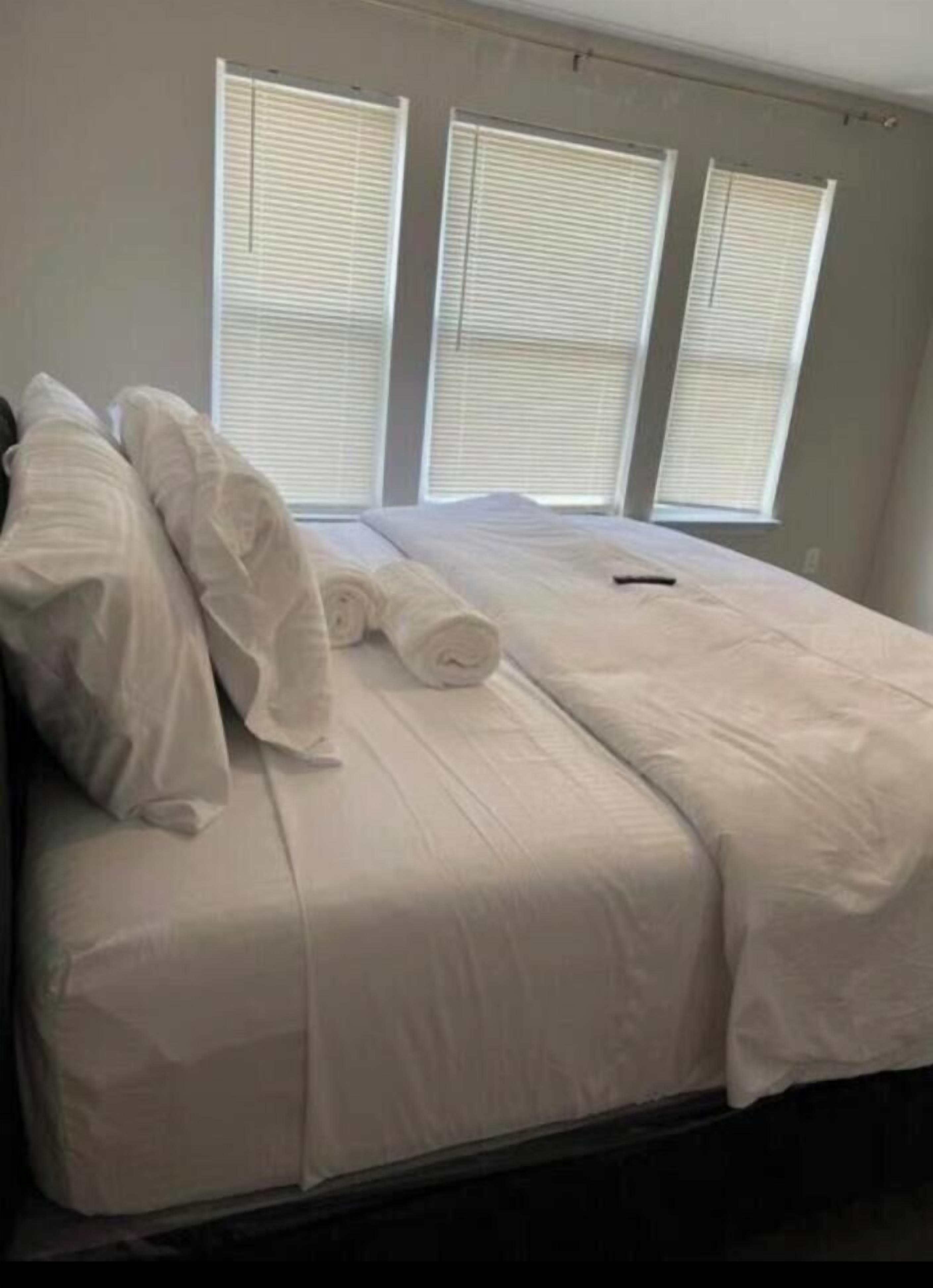 A bed with white bedding, a remote control, and three windows with blinds.