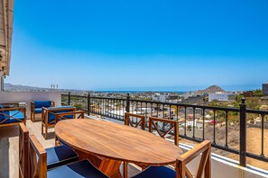 Ein Balkon mit Holztisch und blauen Sitzgelegenheiten, mit Blick auf eine Küstenstadt mit Gebäuden und einem Berg in der Ferne.