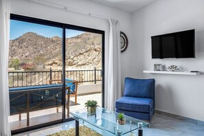 Ein modernes Wohnzimmer mit Glastisch, blauem Sofa, Wand-Fernseher und einem Balkon mit Blick auf eine Berglandschaft.