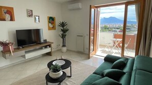 Una sala de estar con un sofá verde, un televisor sobre un mueble bajo, una mesita con una planta y vistas a un balcón con mobiliario exterior.