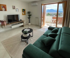 A living room with a green sofa, a TV on a stand, a small table with a plant, and a view of a balcony with outdoor furniture.