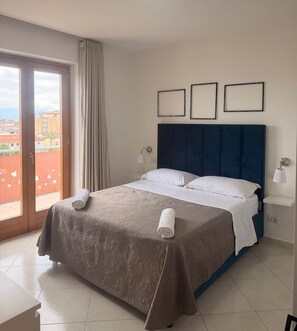 Una camera d'albergo con un letto, due abat-jour, una finestra con tende e vista sugli edifici.