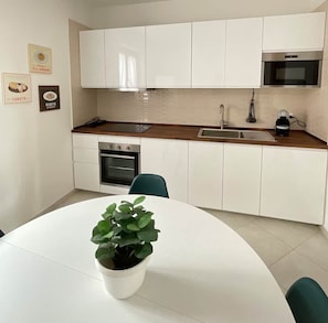A modern kitchen with white cabinets, a microwave, an oven, and a sink. There's a potted plant on the dining table.