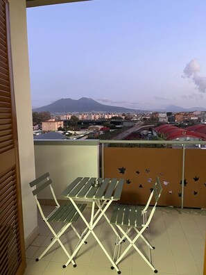 Un balcó amb taula i cadires, una vista d'un paisatge urbà amb edificis i una muntanya, i una paret marró amb adhesius.