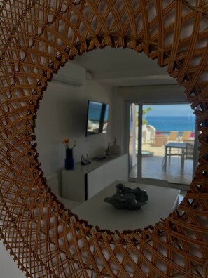 Ein Zimmer mit einem großen Spiegel, einem Flachbildfernseher, einem Essbereich mit Tisch und Stühlen und Meerblick durch eine Glastür.