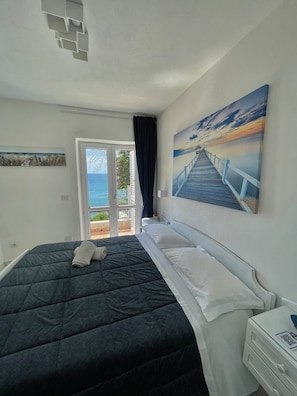 A bedroom with a bed, a painting of a pier, a door leading to a balcony, and a window. - SEA VIEW apartment CAVA dell’ISOLA (Forio)• Elowen (Forio)