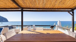 A covered outdoor deck with a wooden table and chairs, overlooking the sea.