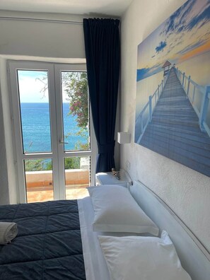 Une chambre d’hôtel avec un lit, un tableau représentant une jetée, une vue sur la mer et un balcon donnant sur la mer.
