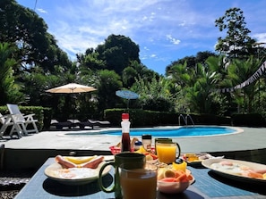 A poolside breakfast setup with a table, chairs, and a view of a swimming pool surrounded by lush greenery. - Casas New Caribe Point (Cahuita)